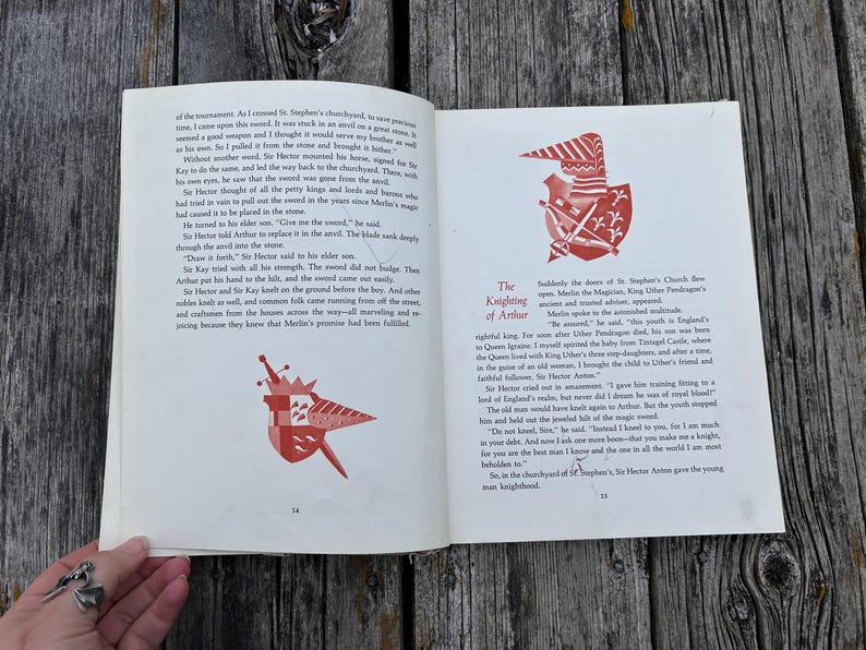 Może przedstawiać: Otwarta książka z czarnym tekstem na białych stronach, zawierająca dwie ilustracje czerwono-białych tarcz herbowych. Lewa strona jest oznaczona numerem 14, a prawa numerem 15, z widocznym tytułem The Knighting of Arthur.