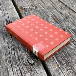 May include: A hardcover book with a red cover featuring a repeating pattern of small, light-colored circular dots. The spine has a white library-style label near the bottom, and the book is positioned at an angle on a weathered wooden surface.