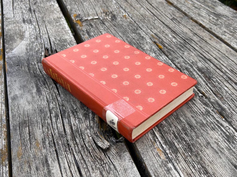 May include: A hardcover book with a red cover featuring a repeating pattern of small, light-colored circular dots. The spine has a white library-style label near the bottom, and the book is positioned at an angle on a weathered wooden surface.