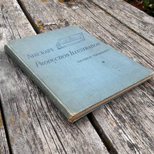 Pu&ograve; includere: Un libro con copertina rigida azzurra intitolato Aircraft Production Illustration di George Tharratt. La copertina presenta un disegno al tratto blu scuro di un componente aeronautico e il titolo stampato in un carattere serif blu scuro.