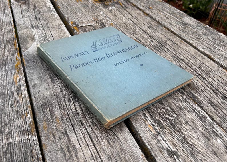 Pu&ograve; includere: Un libro con copertina rigida azzurra intitolato Aircraft Production Illustration di George Tharratt. La copertina presenta un disegno al tratto blu scuro di un componente aeronautico e il titolo stampato in un carattere serif blu scuro.