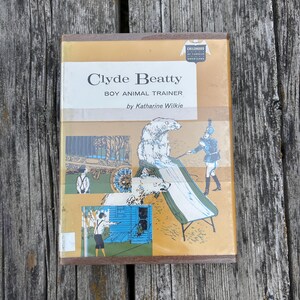 May include: A hardback book titled Clyde Beatty Boy Animal Trainer by Katharine Wilkie. The tan cover features illustrations of a polar bear on a slide, a trainer, and a circus wagon. A Childhood of Famous Americans logo is in the top corner.