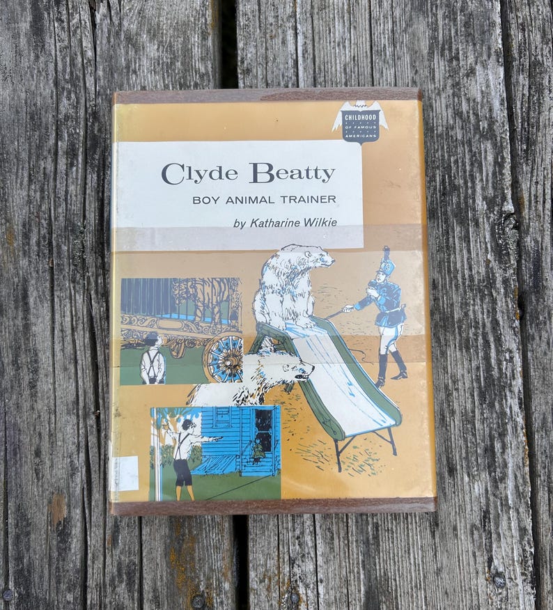 May include: A hardback book titled Clyde Beatty Boy Animal Trainer by Katharine Wilkie. The tan cover features illustrations of a polar bear on a slide, a trainer, and a circus wagon. A Childhood of Famous Americans logo is in the top corner.