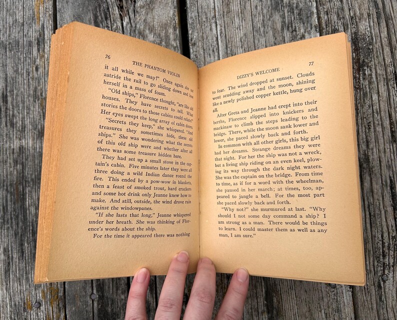 K&ouml;nnte beinhalten: Ein aufgeschlagenes altes Buch mit gealterten, vergilbten Seiten und schwarzem Drucktext. Die linke Seite tr&auml;gt den Titel The Phantom Violin, Seite 76, und die rechte Seite den Titel Dizzy's Welcome, Seite 77. Das Buch zeigt zwei Textspalten.