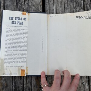 May include: An open hardcover book titled The Story of Our Flag by Carl Glick and Ollie Rogers. The inside front cover features a printed summary, while the opposite page is blank with the word DISCARDED stamped in black ink at the top.