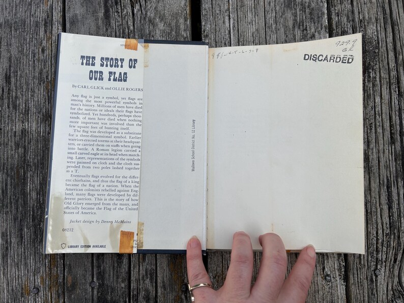 May include: An open hardcover book titled The Story of Our Flag by Carl Glick and Ollie Rogers. The inside front cover features a printed summary, while the opposite page is blank with the word DISCARDED stamped in black ink at the top.