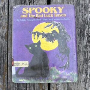 K&ouml;nnte beinhalten: Ein Kinderbuch mit festem Einband mit dem Titel Spooky and the Bad Luck Raven von Natalie Savage Carlson, illustriert von Andrew Glass. Das violette Cover zeigt eine schwarze Katze mit gr&uuml;nen Augen und einen Raben vor einem gelben Mond.