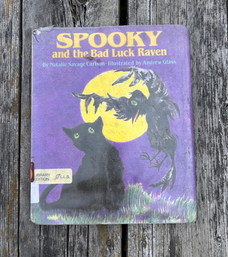 K&ouml;nnte beinhalten: Ein Kinderbuch mit festem Einband mit dem Titel Spooky and the Bad Luck Raven von Natalie Savage Carlson, illustriert von Andrew Glass. Das violette Cover zeigt eine schwarze Katze mit gr&uuml;nen Augen und einen Raben vor einem gelben Mond.