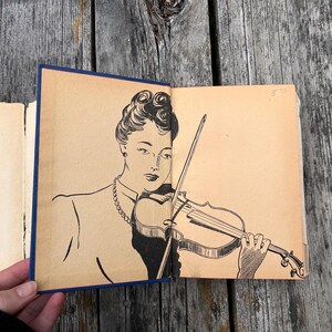 May include: A vintage book with a tan endpaper illustration featuring a black ink line drawing of a person with an updo hairstyle wearing a necklace and playing a violin with a bow. The book has a dark blue spine and aged paper edges.