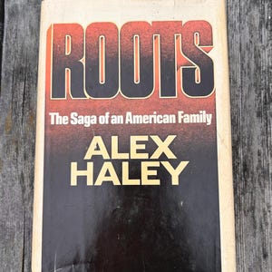 K&ouml;nnte beinhalten: Ein gebundenes Buch mit dem Titel Roots: The Saga of an American Family von Alex Haley. Der Einband zeigt den Titel in fetten, schwarzen Buchstaben vor einem rot-orangen Farbverlauf, mit dem Namen des Autors in gro&szlig;en, gelben Gro&szlig;buchstaben.