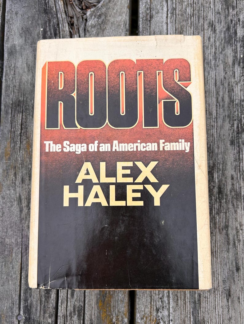 K&ouml;nnte beinhalten: Ein gebundenes Buch mit dem Titel Roots: The Saga of an American Family von Alex Haley. Der Einband zeigt den Titel in fetten, schwarzen Buchstaben vor einem rot-orangen Farbverlauf, mit dem Namen des Autors in gro&szlig;en, gelben Gro&szlig;buchstaben.