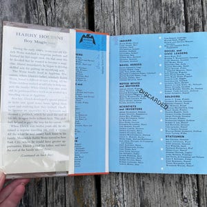 May include: An open book with a light blue dust jacket featuring a list of historical figures categorized by roles such as Indians, Naval Heroes, and Scientists. The left side contains a biography of Harry Houdini, and the word DISCARDED is stamped.