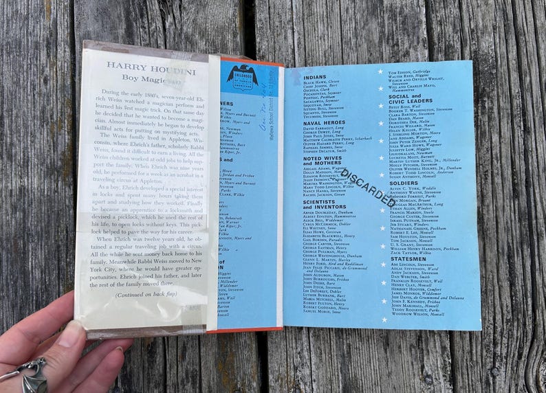May include: An open book with a light blue dust jacket featuring a list of historical figures categorized by roles such as Indians, Naval Heroes, and Scientists. The left side contains a biography of Harry Houdini, and the word DISCARDED is stamped.
