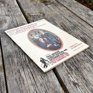 K&ouml;nnte beinhalten: Ein Notenheft f&uuml;r Butch Cassidy and the Sundance Kid mit cremefarbenem Einband, einer zentralen ovalen Illustration von drei Personen und rotem sowie schwarzem Text, der die Namen Paul Newman und Robert Redford enth&auml;lt.