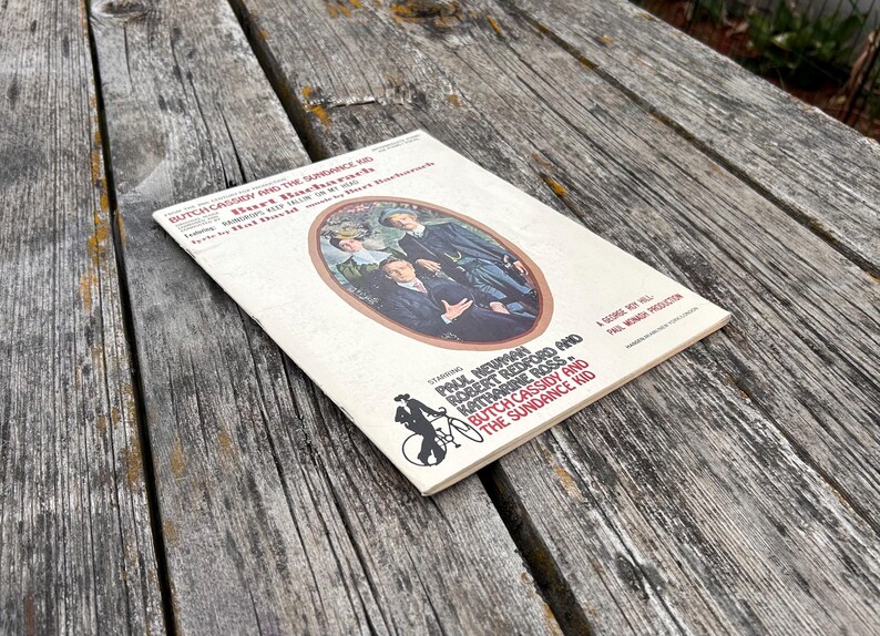 K&ouml;nnte beinhalten: Ein Notenheft f&uuml;r Butch Cassidy and the Sundance Kid mit cremefarbenem Einband, einer zentralen ovalen Illustration von drei Personen und rotem sowie schwarzem Text, der die Namen Paul Newman und Robert Redford enth&auml;lt.