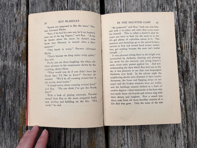 May include: An open vintage book titled Roy Blakeley In The Haunted Camp with pages 58 and 59 visible. The text is printed in black ink on off-white paper. The left page features dialogue between characters, while the right page continues the narrative.