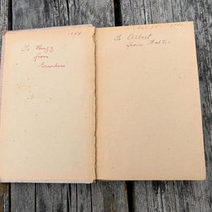 K&ouml;nnte beinhalten: Ein aufgeschlagenes altes Buch mit gealterten, hellbraunen Seiten. Die linke Seite zeigt handgeschriebenen Text in roter Tinte mit 'To Buzz from Grandma' und '1956'. Die rechte Seite enth&auml;lt handgeschriebenen Text in schwarzer Tinte mit 'Dec 25 - 1950' und 'To Albert from Walter'.