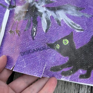 K&ouml;nnte beinhalten: Ein violetter, strukturierter Druck mit einer schwarzen Kr&auml;he mit ausgebreiteten Fl&uuml;geln und einer schwarzen Katze mit leuchtend gr&uuml;nen Augen. In der oberen Ecke ist eine gelbe Kreisform zu sehen. Das Wort DISCARDED ist schwarz neben dem Kopf der Katze gedruckt.