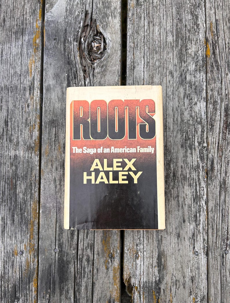 K&ouml;nnte beinhalten: Ein gebundenes Buch mit dem Titel Roots: The Saga of an American Family von Alex Haley. Der Buchumschlag zeigt eine kr&auml;ftige schwarze und rote Schrift auf schwarzem Hintergrund, eingerahmt von einem cremefarbenen Rand auf der Vorderseite.