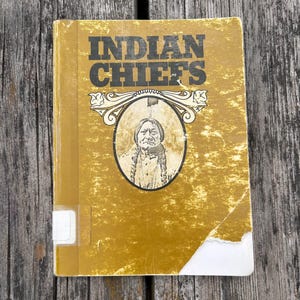 Może przedstawiać: Musztardowa książka w twardej oprawie zatytułowana INDIAN CHIEFS z centralną owalną ilustracją przedstawiającą osobę z warkoczami. Okładka nosi ślady zużycia, w tym podarty dolny r&oacute;g i białą etykietę biblioteczną na grzbiecie.