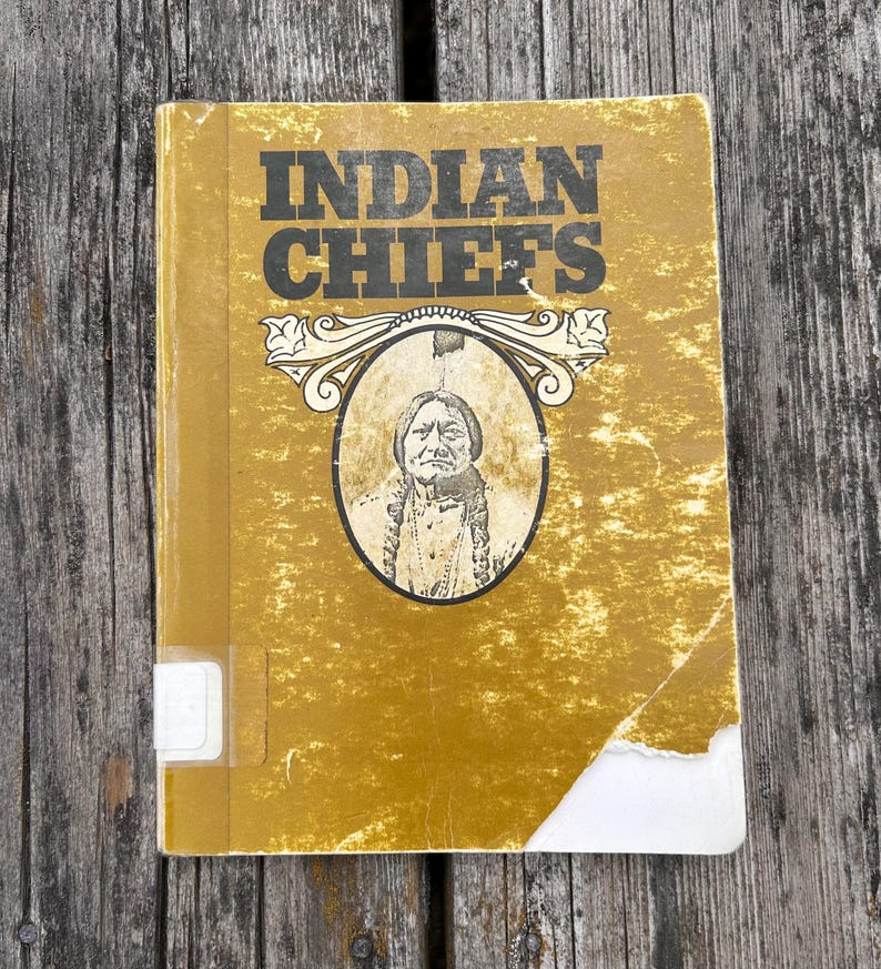 Może przedstawiać: Musztardowa książka w twardej oprawie zatytułowana INDIAN CHIEFS z centralną owalną ilustracją przedstawiającą osobę z warkoczami. Okładka nosi ślady zużycia, w tym podarty dolny r&oacute;g i białą etykietę biblioteczną na grzbiecie.