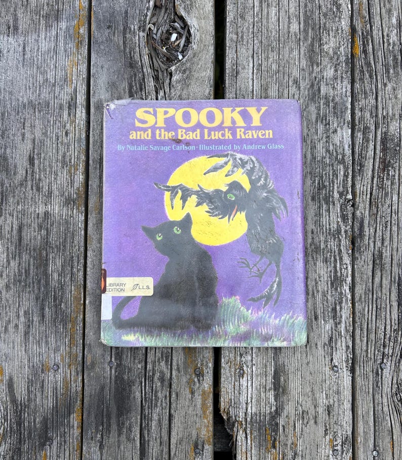 K&ouml;nnte beinhalten: Ein gebundenes Buch mit dem Titel Spooky and the Bad Luck Raven von Natalie Savage Carlson, illustriert von Andrew Glass. Der violette Einband zeigt eine schwarze Katze mit gr&uuml;nen Augen, einen gro&szlig;en gelben Mond und einen schwarzen Raben.