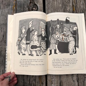 Puede incluir: Un libro abierto con ilustraciones en blanco y negro de varios personajes, incluyendo un esqueleto y un gato, reunidos alrededor de un gran caldero. El texto dice: Ms. McWee has strange friends who come to visit. They bring big books of magic and cases filled with magic charms. Witches hold special meetings where they share their magic secrets. My mother says, Those people are members of Ms. McWee's reading and music club.