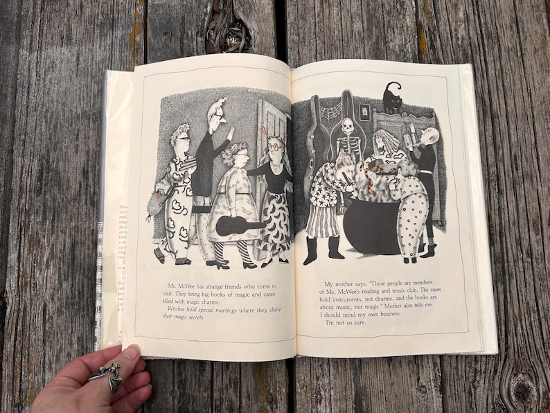 Puede incluir: Un libro abierto con ilustraciones en blanco y negro de varios personajes, incluyendo un esqueleto y un gato, reunidos alrededor de un gran caldero. El texto dice: Ms. McWee has strange friends who come to visit. They bring big books of magic and cases filled with magic charms. Witches hold special meetings where they share their magic secrets. My mother says, Those people are members of Ms. McWee's reading and music club.