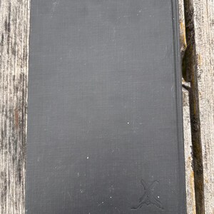 Pu&ograve; includere: Un libro con copertina rigida rettangolare rivestito in tessuto nero opaco. La copertina presenta un piccolo logo stilizzato in rilievo nell'angolo in basso a destra, che ricorda un paio di spade incrociate o un emblema decorativo grigio scuro.