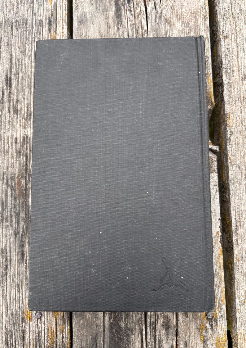 Pu&ograve; includere: Un libro con copertina rigida rettangolare rivestito in tessuto nero opaco. La copertina presenta un piccolo logo stilizzato in rilievo nell'angolo in basso a destra, che ricorda un paio di spade incrociate o un emblema decorativo grigio scuro.