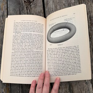Peut inclure: Livre ouvert avec du texte et un diagramme d'un tore. Les pages sont blanc cass&eacute; et le livre est pos&eacute; sur une surface en bois. Le texte traite de physique et de la th&eacute;orie des cordes. Le diagramme illustre des dimensions suppl&eacute;mentaires.