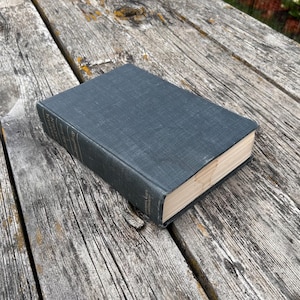 May include: A hardcover book with a dark charcoal gray fabric cover and a textured weave. The spine features faint, partially visible white lettering. The pages are cream-colored and have a smooth, clean edge, showing a rectangular shape.