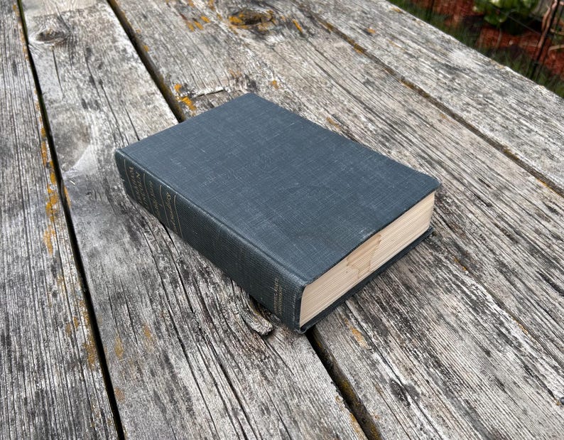 May include: A hardcover book with a dark charcoal gray fabric cover and a textured weave. The spine features faint, partially visible white lettering. The pages are cream-colored and have a smooth, clean edge, showing a rectangular shape.