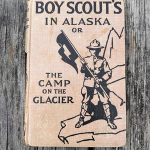 K&ouml;nnte beinhalten: Ein gebundenes Vintage-Buch mit dem Titel BOY SCOUT'S IN ALASKA OR THE CAMP ON THE GLACIER. Der beigefarbene Einband zeigt eine schwarze Tuschezeichnung eines Pfadfinders in Uniform mit einer Flagge und Bergumrissen sowie abgenutzten Kanten.