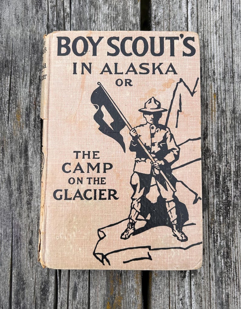 K&ouml;nnte beinhalten: Ein gebundenes Vintage-Buch mit dem Titel BOY SCOUT'S IN ALASKA OR THE CAMP ON THE GLACIER. Der beigefarbene Einband zeigt eine schwarze Tuschezeichnung eines Pfadfinders in Uniform mit einer Flagge und Bergumrissen sowie abgenutzten Kanten.