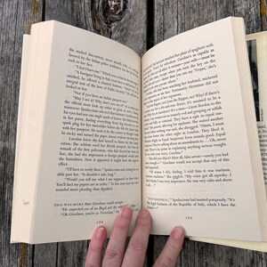 May include: An open paperback book displaying pages 194 and 195 of a novel. The pages contain printed black text in English, featuring dialogue and narrative paragraphs. The book has off-white paper and is held open to reveal the printed content.