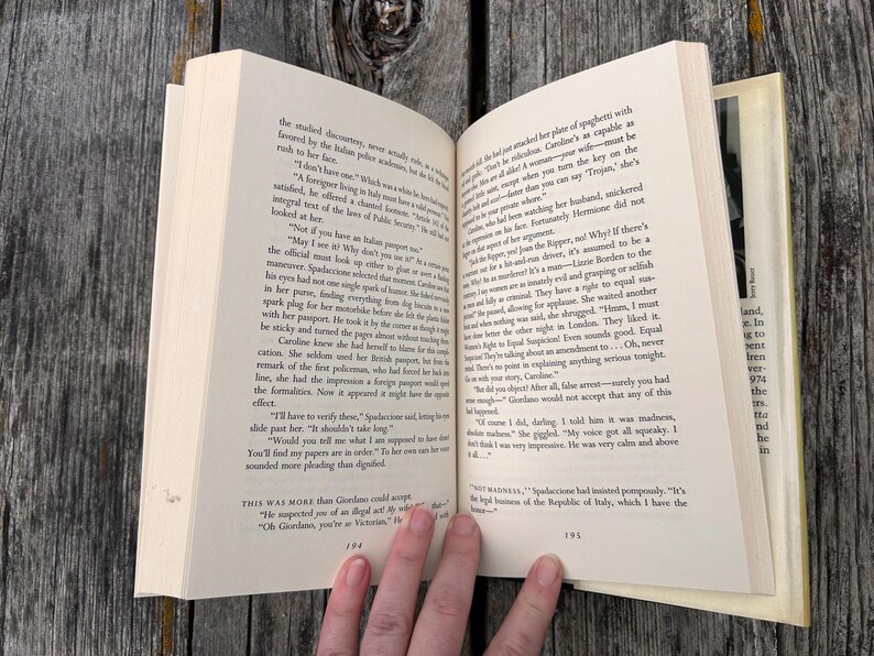 May include: An open paperback book displaying pages 194 and 195 of a novel. The pages contain printed black text in English, featuring dialogue and narrative paragraphs. The book has off-white paper and is held open to reveal the printed content.