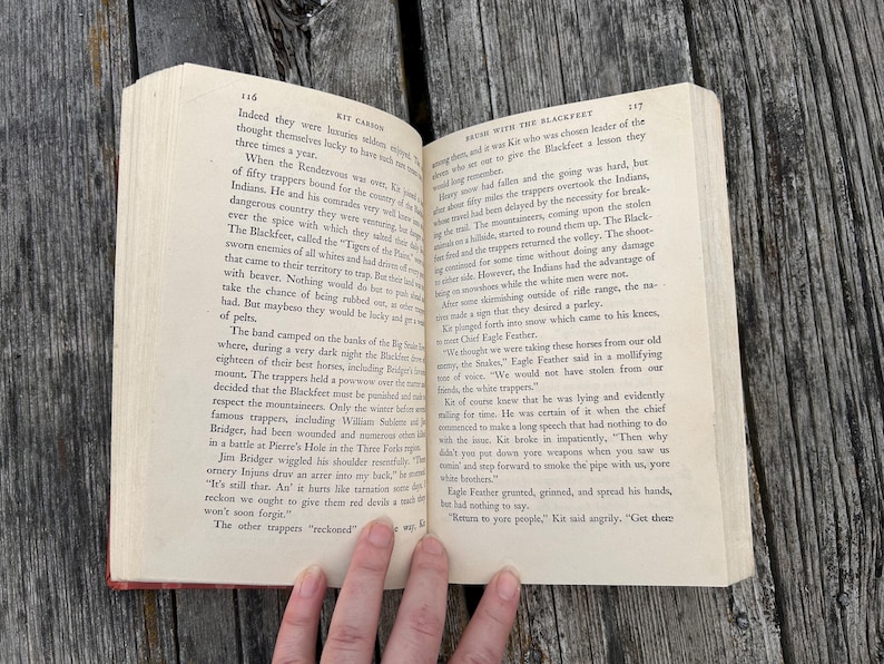 May include: An open hardcover book with cream-colored pages featuring black printed text. The left page is numbered 116 and the right page is numbered 117, with the chapter title Brush with the Blackfeet visible at the top.