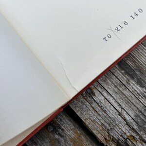 May include: An open book with cream-colored pages featuring a red cover edge. The top right corner of the page displays the printed numbers 70, 21, 6, and 140, with a faint diagonal line crossing through the number 21 in black ink.