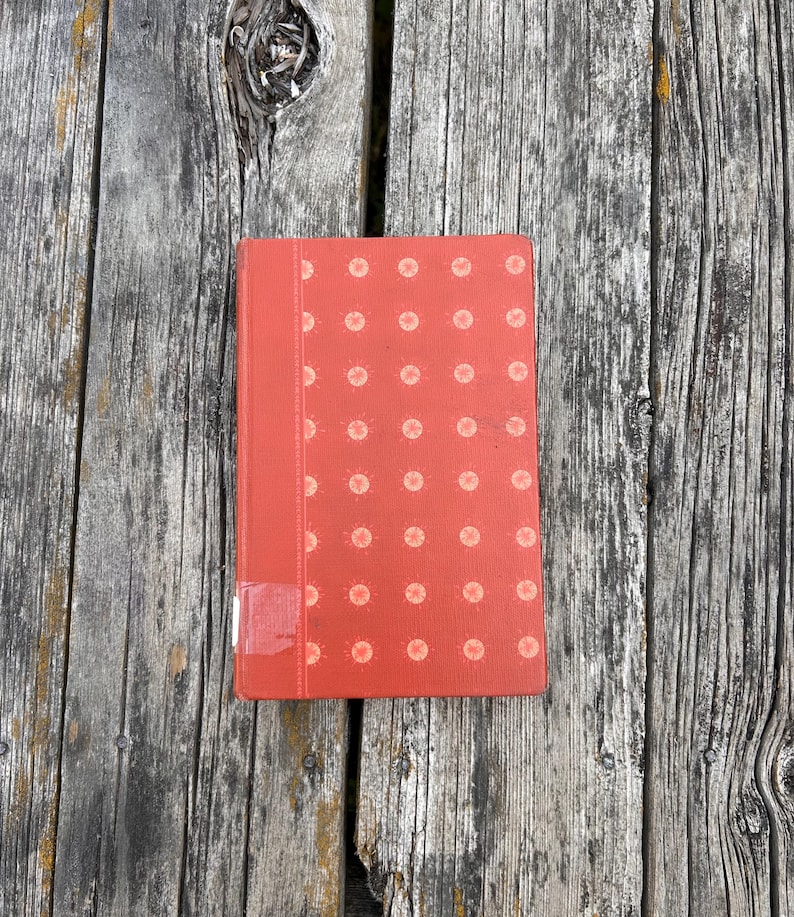May include: A rectangular hardcover book with a vibrant orange cover featuring a repeating pattern of light-colored circular motifs. A vertical strip runs along the left side, and a small white rectangular label is attached to the bottom corner.