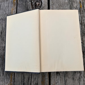May include: An open hardcover book with blank, off-white pages. The book is positioned so that the spine is visible on the left side, showing a dark cover, while the interior pages are completely empty and feature a smooth, matte texture.