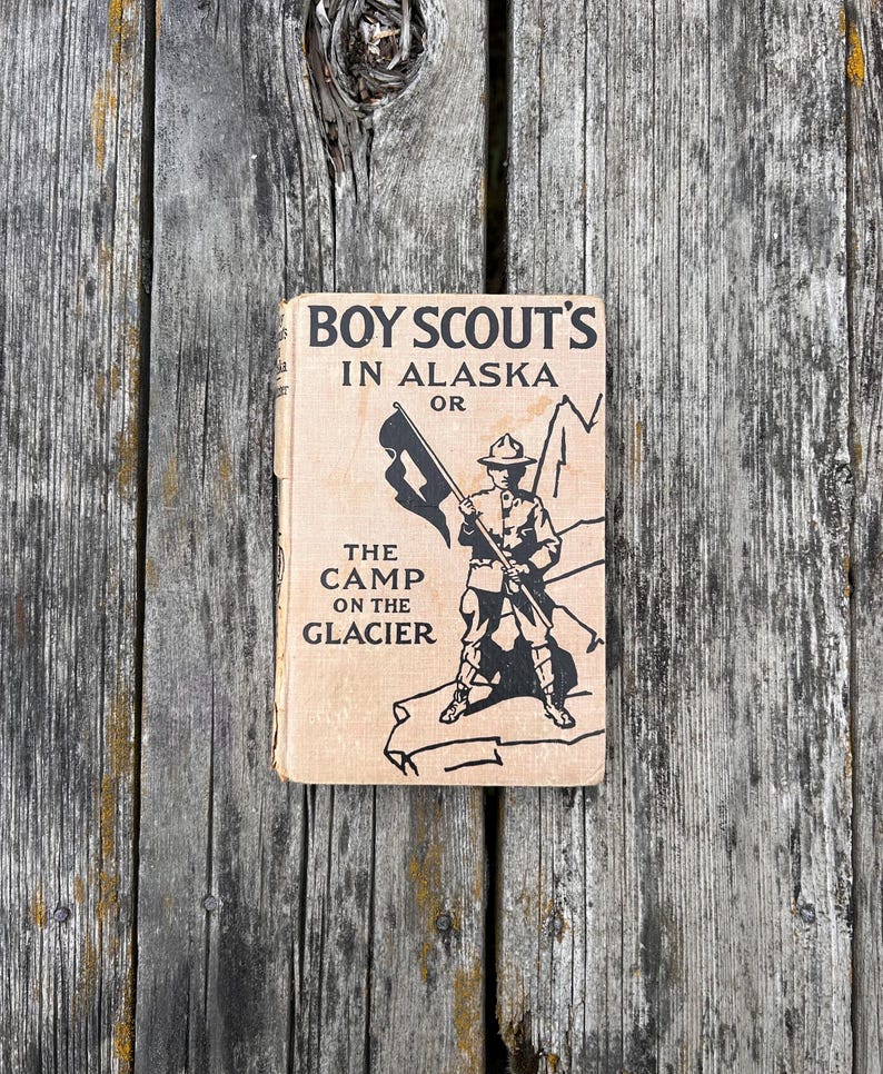 K&ouml;nnte beinhalten: Ein gebundenes Vintage-Buch mit dem Titel Boy Scout's in Alaska or The Camp on the Glacier. Der beigefarbene Einband zeigt eine schwarze Tuschezeichnung eines Pfadfinders mit einer Flagge auf einem Gletscher. Der Text ist in Serifenschrift.