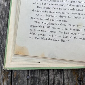 K&ouml;nnte beinhalten: Ein aufgeschlagenes altes Buch mit hellgr&uuml;nem Einband zeigt eine Seite mit gedrucktem Text aus einer Geschichte &uuml;ber Hiawatha und Mudjekeewis. Das Papier ist cremefarben mit sichtbaren gelb-braunen Wasserflecken am unteren Seitenrand.