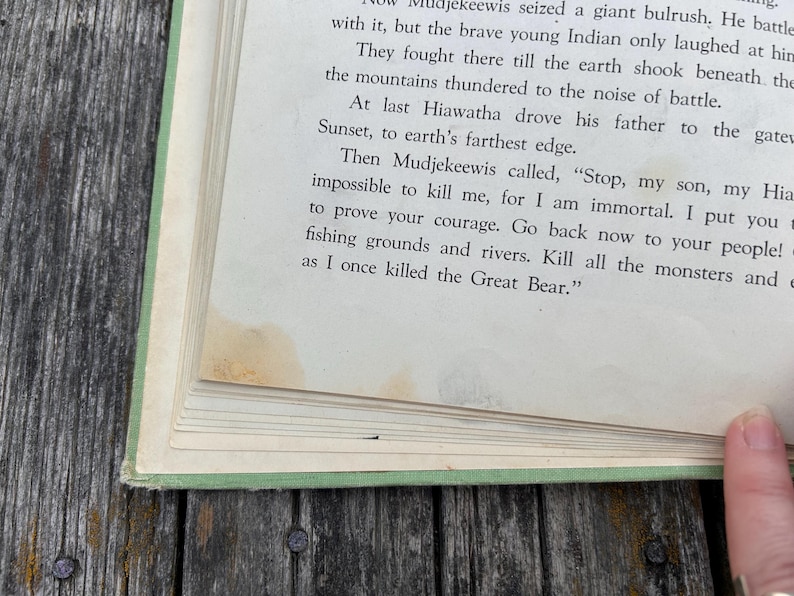 K&ouml;nnte beinhalten: Ein aufgeschlagenes altes Buch mit hellgr&uuml;nem Einband zeigt eine Seite mit gedrucktem Text aus einer Geschichte &uuml;ber Hiawatha und Mudjekeewis. Das Papier ist cremefarben mit sichtbaren gelb-braunen Wasserflecken am unteren Seitenrand.