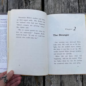 May include: An open book with off-white pages featuring black printed text. The left page shows page number 14 with text about Helen's parents. The right page displays Chapter 2 titled The Stranger and page number 15 with text about Helen waking up.