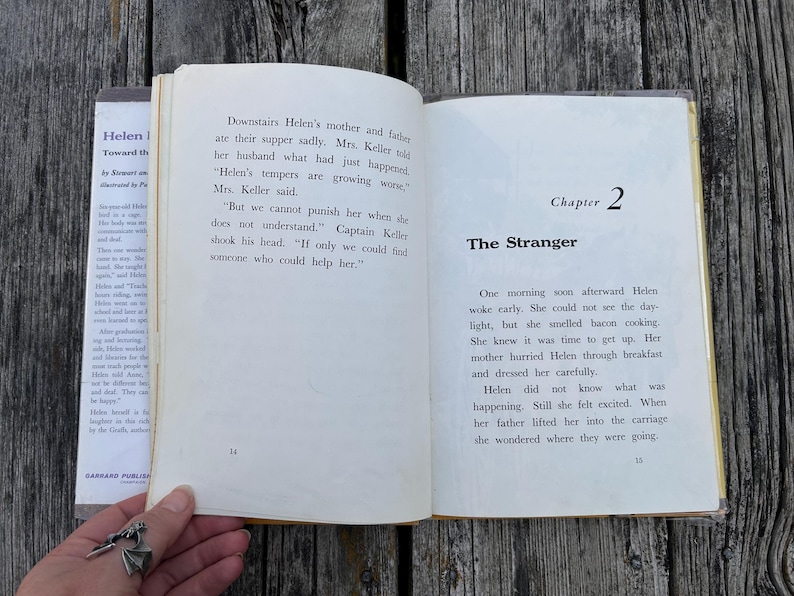 May include: An open book with off-white pages featuring black printed text. The left page shows page number 14 with text about Helen's parents. The right page displays Chapter 2 titled The Stranger and page number 15 with text about Helen waking up.