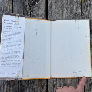 May include: An open hardback book titled Helen Keller: Toward the Light by Stewart and Polly Anne Graff, illustrated by Paul Frame. The dust jacket flap features printed biographical text, and the inner page shows a small stamp at the bottom.