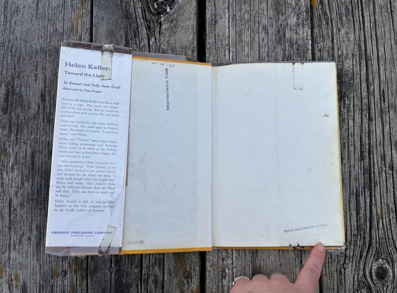 May include: An open hardback book titled Helen Keller: Toward the Light by Stewart and Polly Anne Graff, illustrated by Paul Frame. The dust jacket flap features printed biographical text, and the inner page shows a small stamp at the bottom.