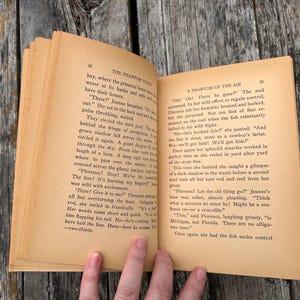 K&ouml;nnte beinhalten: Ein aufgeschlagenes altes Buch mit gealterten, hellbraunen Seiten und schwarzem Drucktext. Die linke Seite tr&auml;gt den Titel The Phantom Violin und die rechte Seite A Phantom of the Air. Das Papier weist deutliche Spuren von Vergilbung auf.