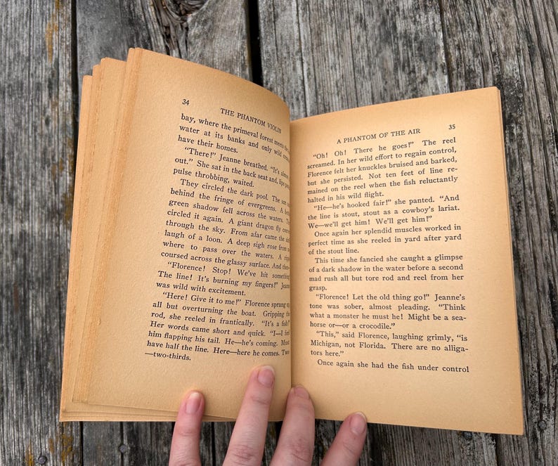 K&ouml;nnte beinhalten: Ein aufgeschlagenes altes Buch mit gealterten, hellbraunen Seiten und schwarzem Drucktext. Die linke Seite tr&auml;gt den Titel The Phantom Violin und die rechte Seite A Phantom of the Air. Das Papier weist deutliche Spuren von Vergilbung auf.